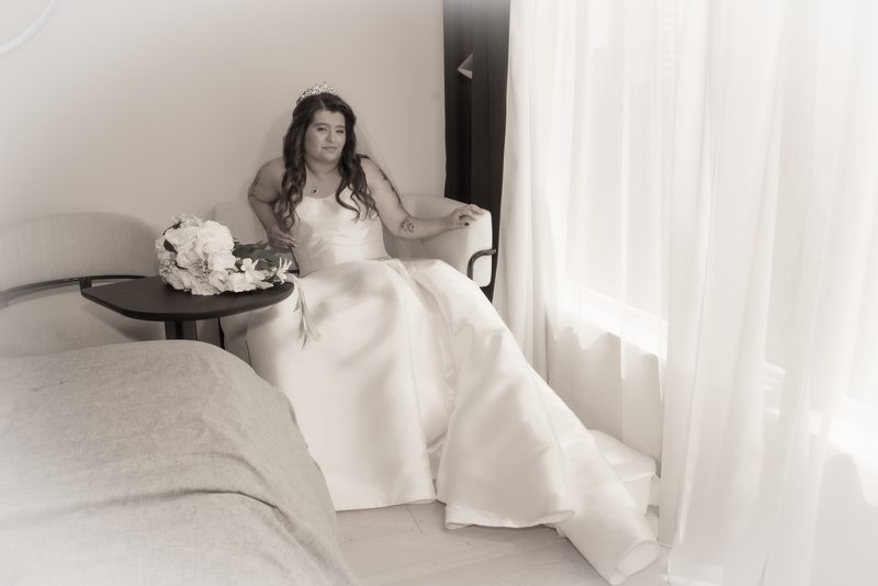 A bride in her wedding gown sits quietly by a window, gazing thoughtfully as light filters through curtains.