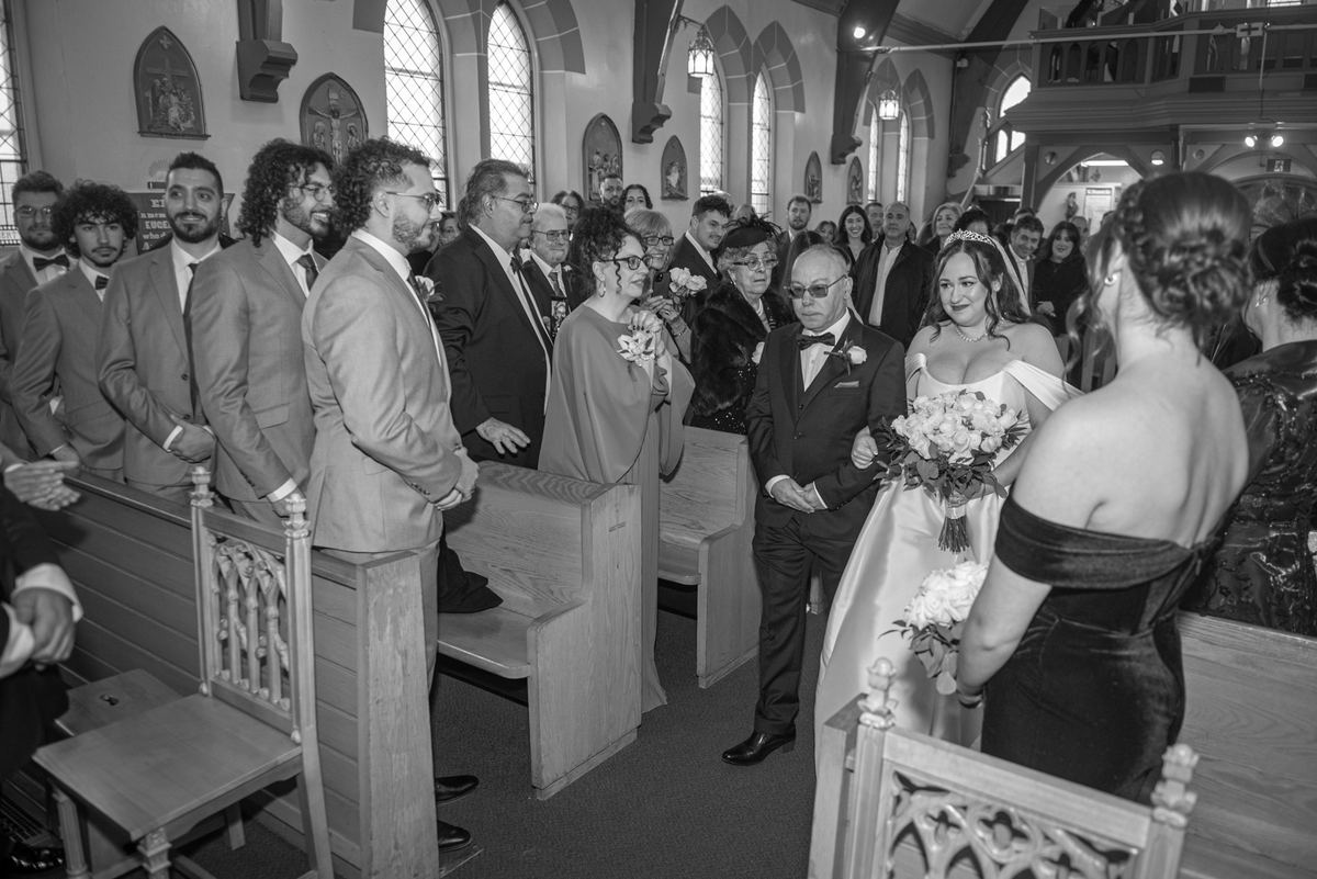 Bride and groom walk down the aisle toward the altar in a church, surrounded by guests in formal attire.