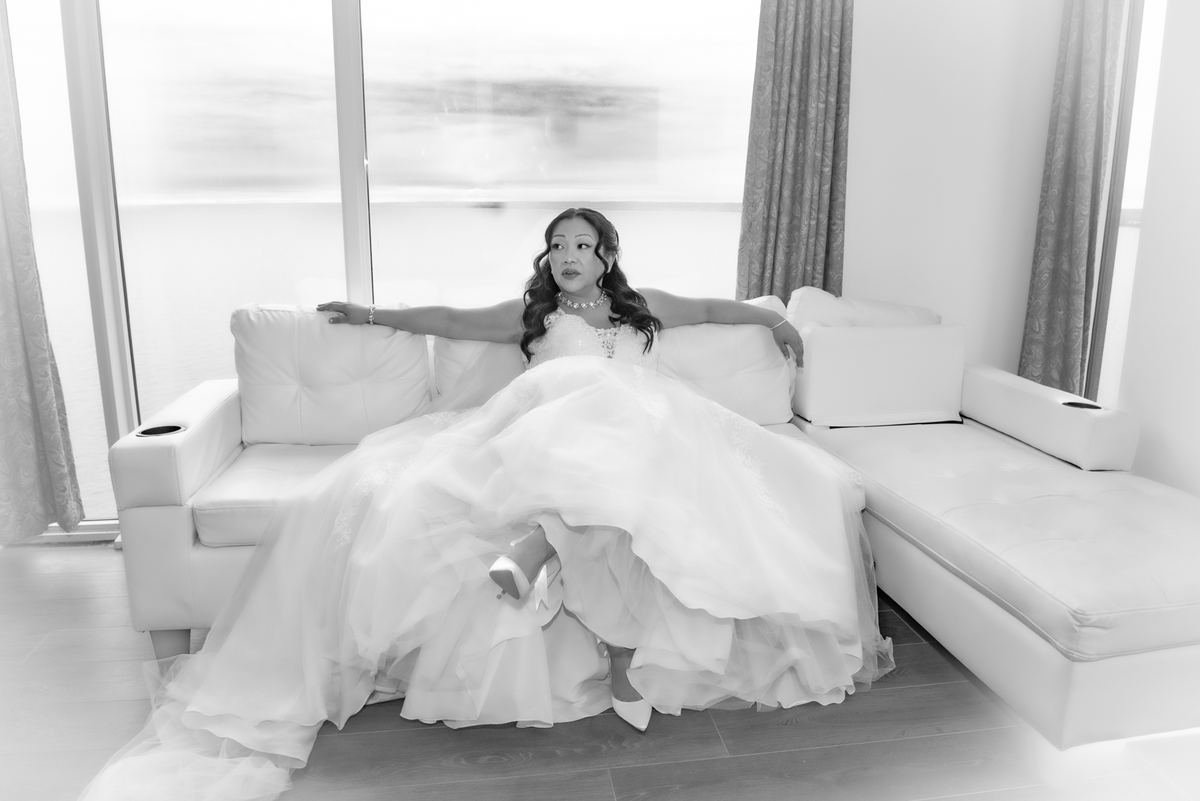 Bride in white gown sits relaxed on modern sofa, arms outstretched, gazing thoughtfully toward camera.