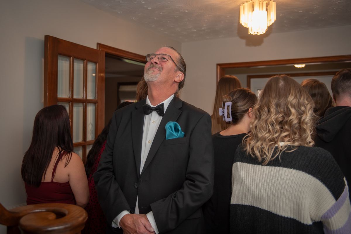 A man in a tuxedo with his eyes closed stands amidst guests at a formal event, illuminated by chandelier light.