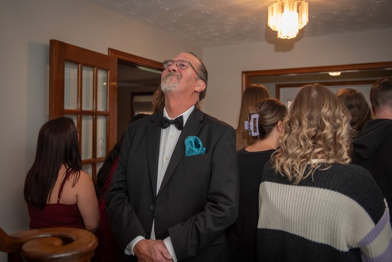 A man in a tuxedo with his eyes closed stands amidst guests at a formal event, illuminated by chandelier light.
