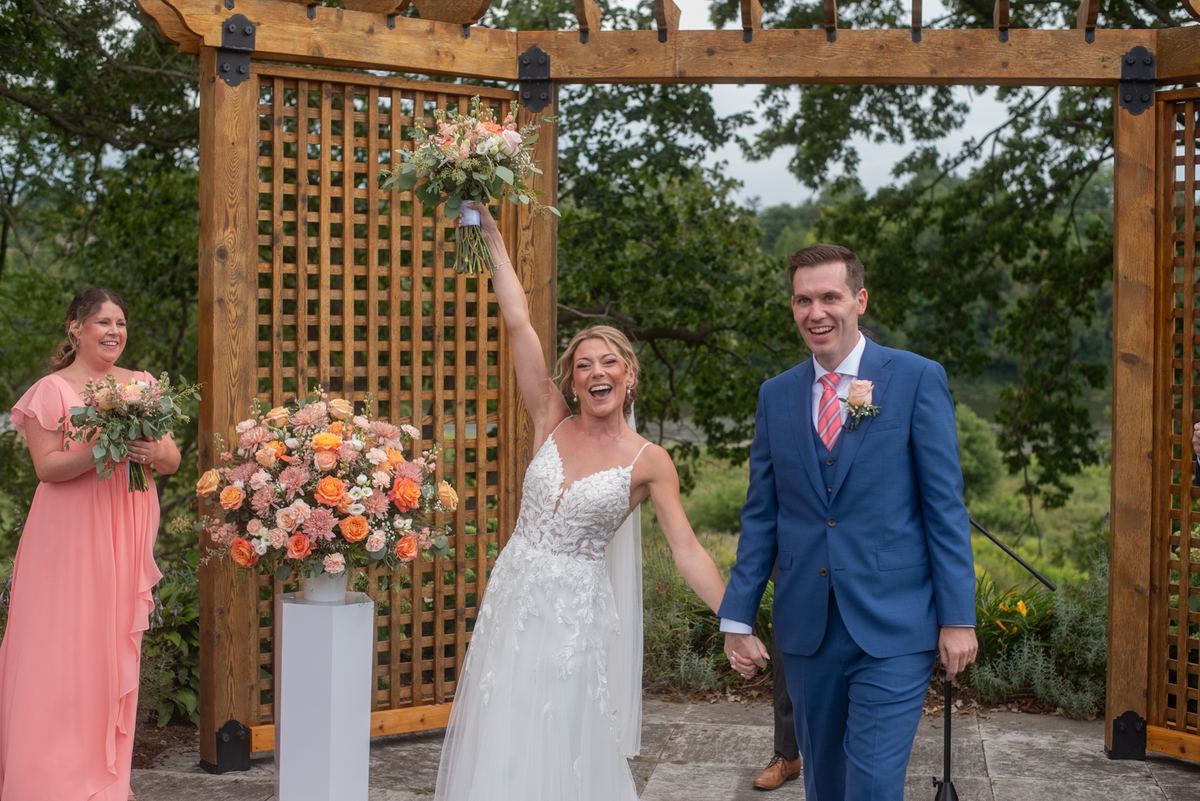 Bride in white lace gown laughs with bouquet raised as groom in blue suit walks hand-in-hand down aisle under wooden arbor.