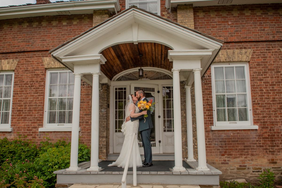 Bride and groom kiss on a porch with white columns and wooden portico, illuminated by soft daylight.