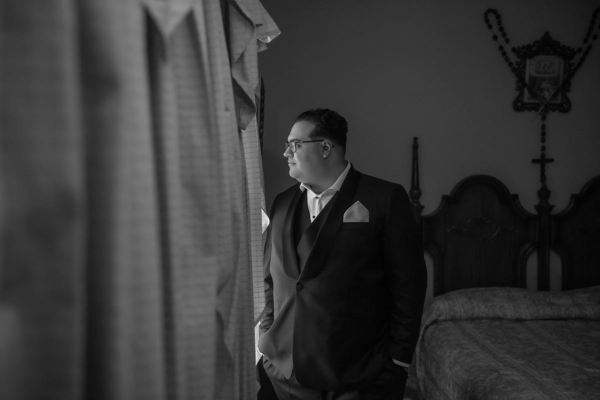 A man in a formal suit stands beside an ornately designed headboard, gazing thoughtfully towards the left of the frame.