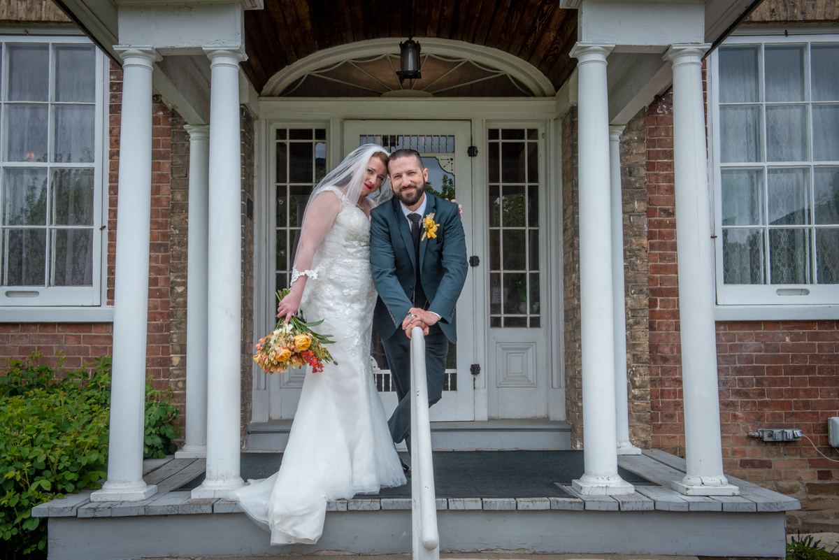 A bride and groom pose affectionately on a porch, the soft light highlighting their smiles and the brick house's columns.