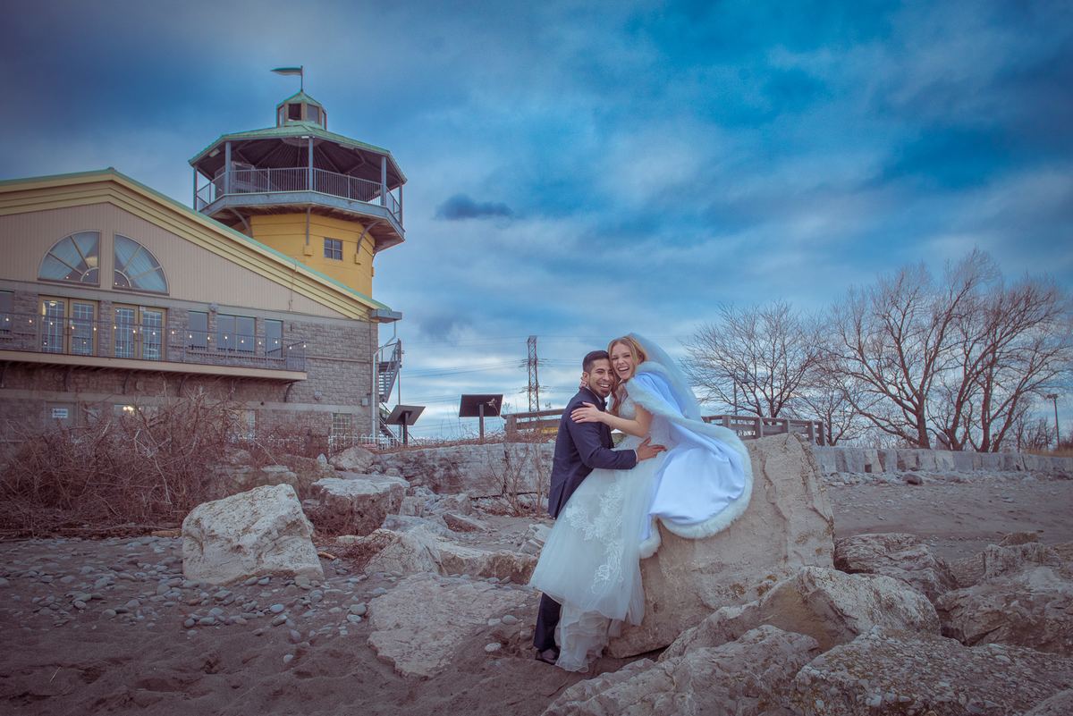 A bride and groom embrace on a rocky shore at dawn, with a cloudy sky and a lighthouse-style building in the background.