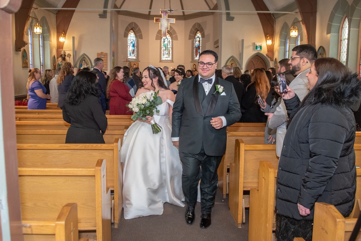 A bride in white and groom in tuxedo walk down the aisle of a church, smiling at each other as guests watch.