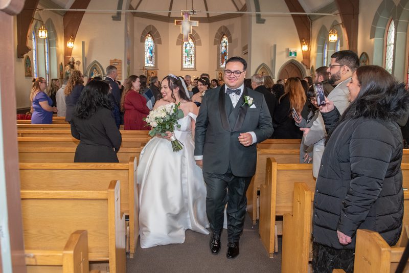 A bride in white and groom in tuxedo walk down the aisle of a church, smiling at each other as guests watch.
