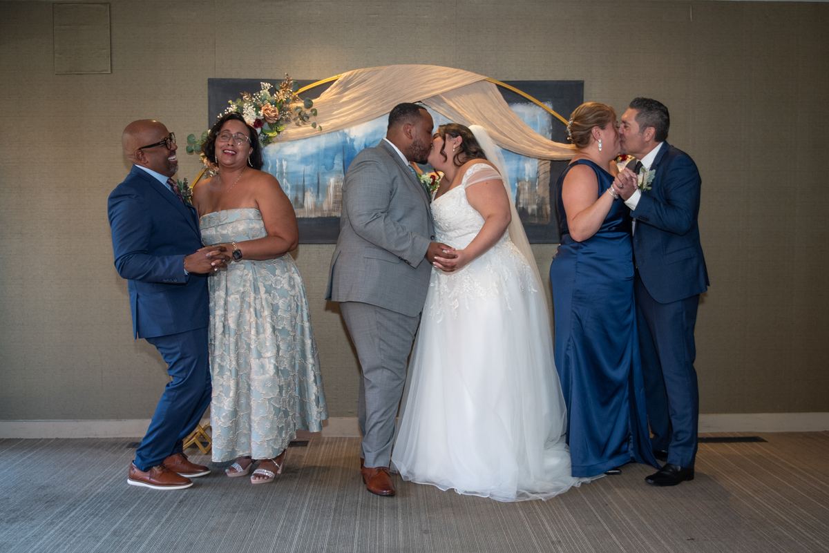 Two couples kiss joyfully, with the bride and groom at center amid smiling family members under an elegant floral backdrop.