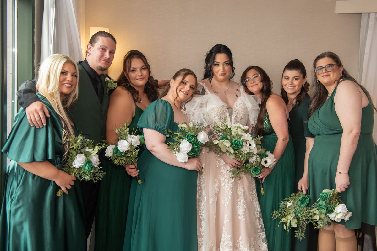 Bride in lace gown and bridal party in emerald green dresses pose with bouquets, illuminated by soft lamp light.