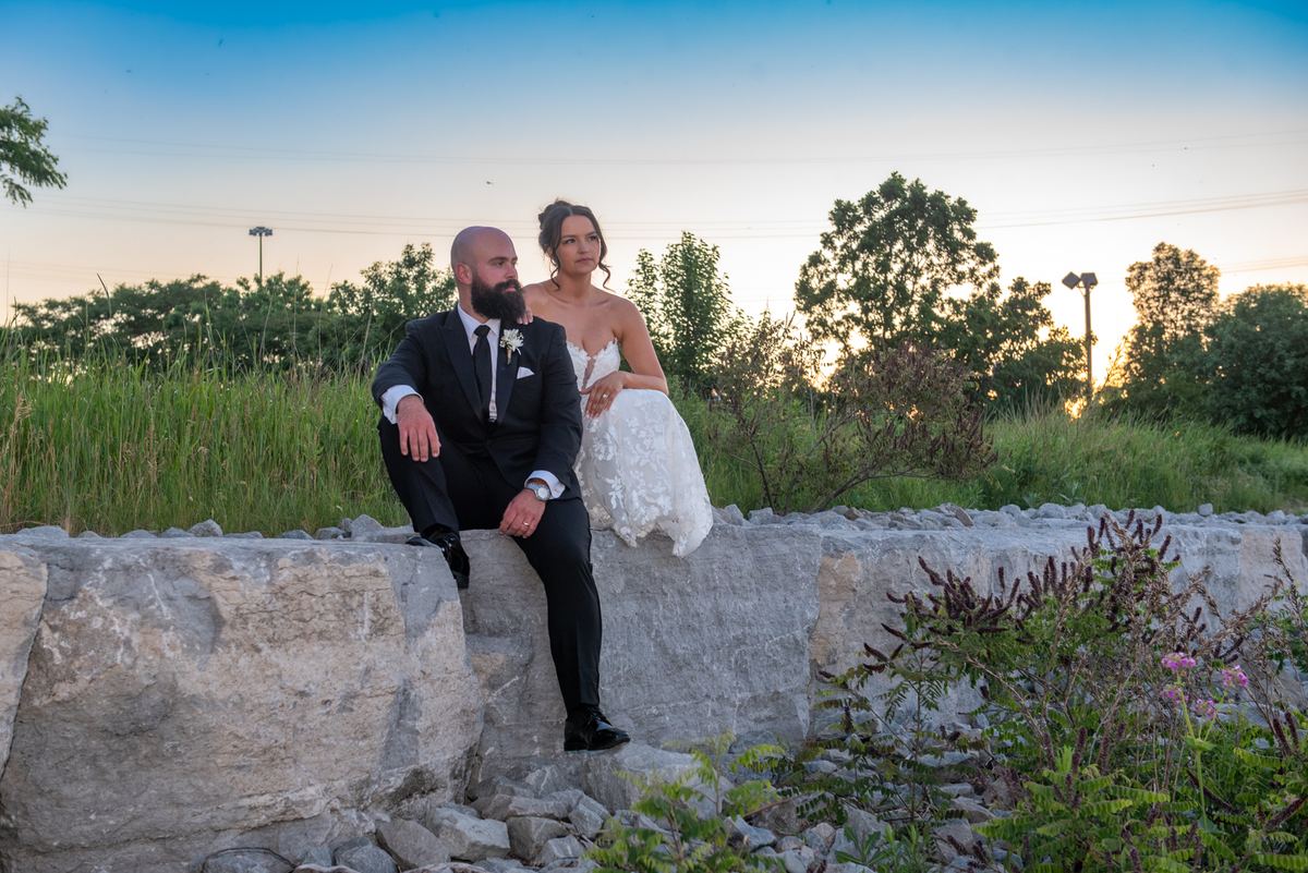 A newlywed couple in formal attire sit contemplatively on a stone wall at sunset, bathed in warm light.