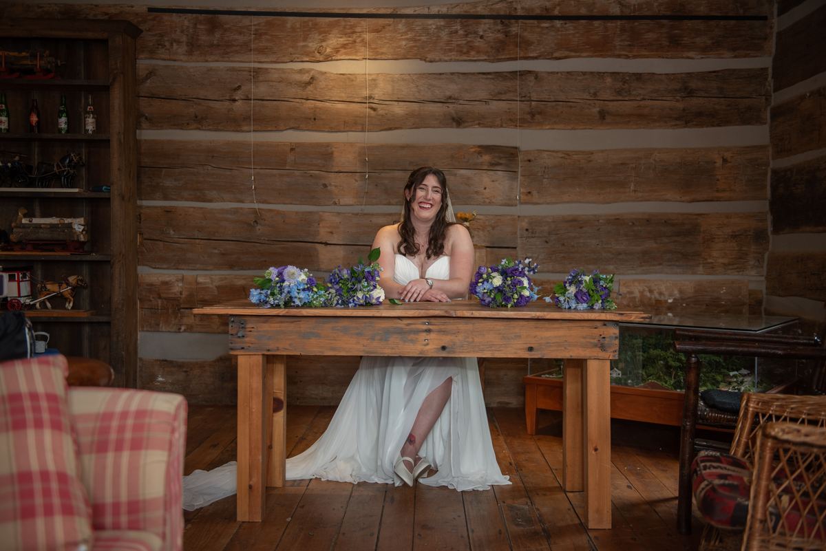 A smiling bride in a white gown sits at a rustic wooden table surrounded by floral arrangements in warm lighting.