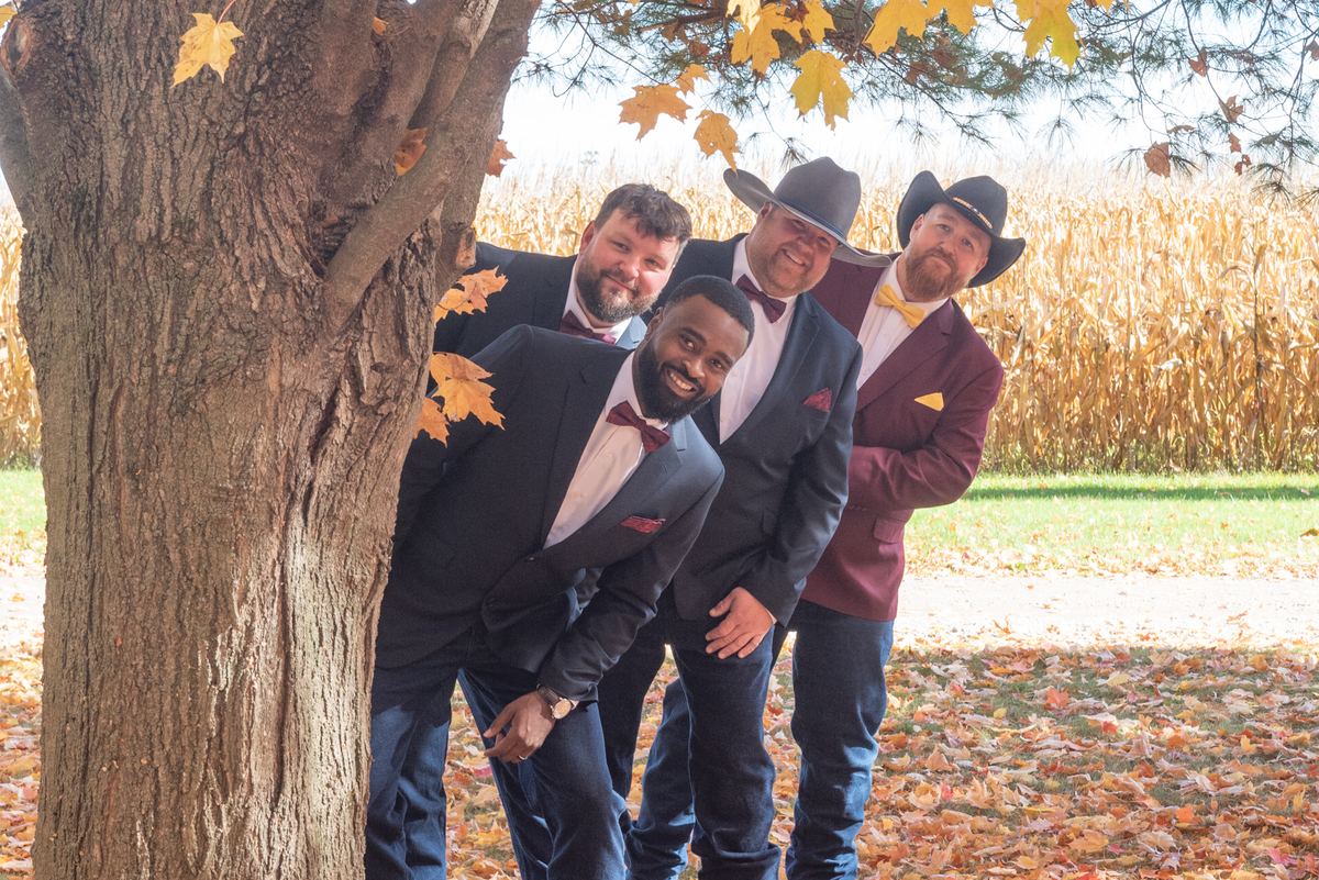 Four men in suits and bow ties, some with cowboy hats, peek behind a large autumn tree near a cornfield.