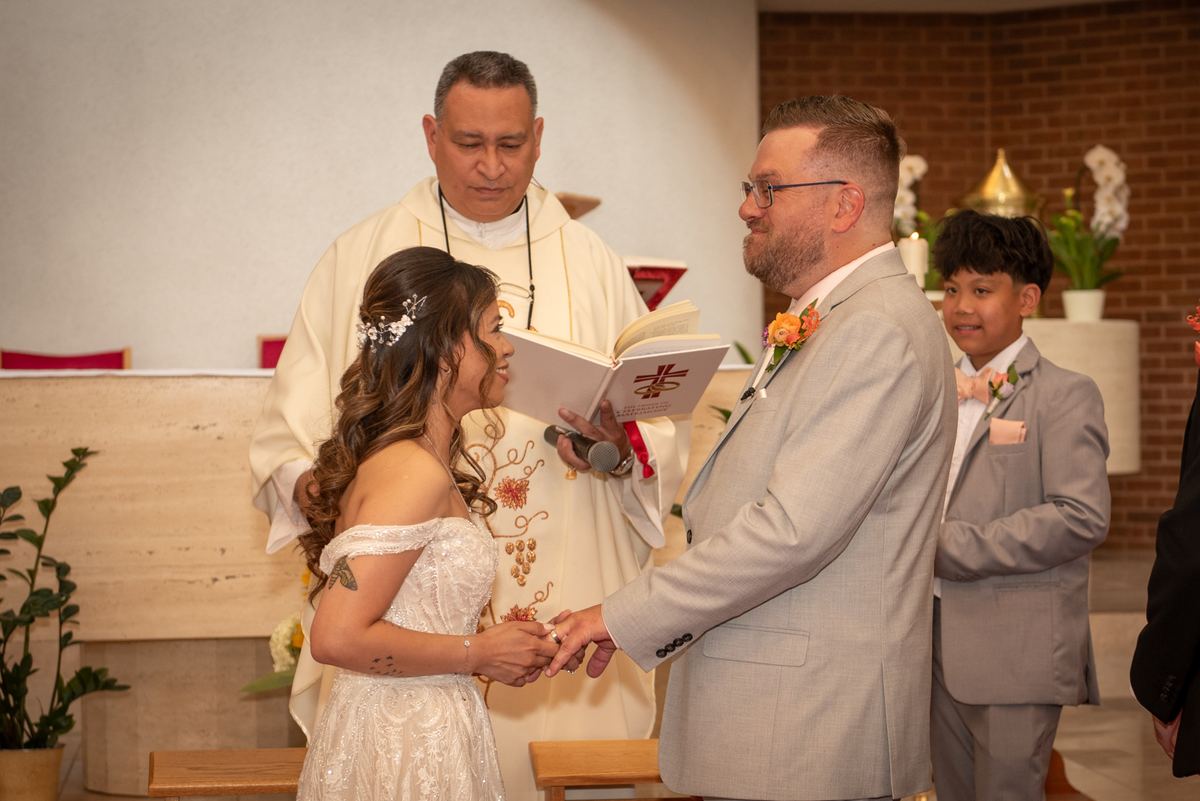 Bride and groom hold hands at altar as priest officiates during wedding ceremony, ring bearer nearby.