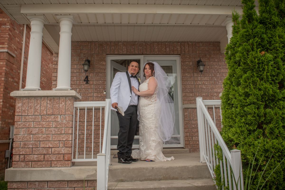 A smiling bride in a lace gown and veil holds hands with her groom in a tuxedo on the front porch steps of a brick home.
