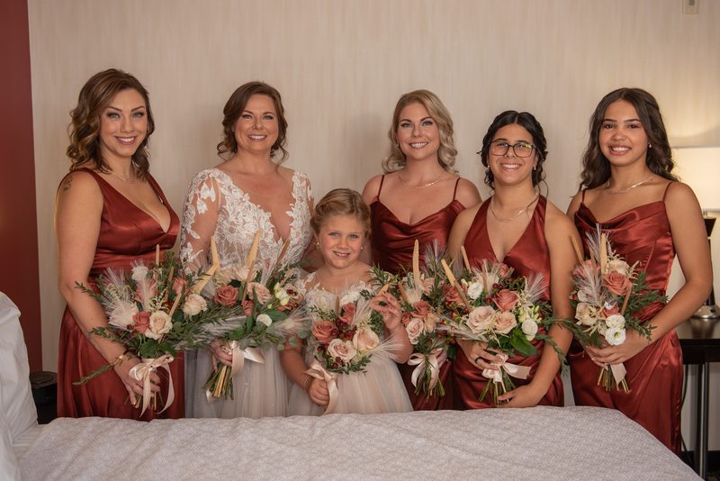 The bride and her smiling bridesmaids hold matching bouquets in a softly lit hotel room, their satin gowns gleaming.