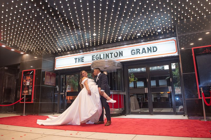 A smiling bride is lifted by her groom on a red carpet outside a theater, surrounded by twinkling lights.