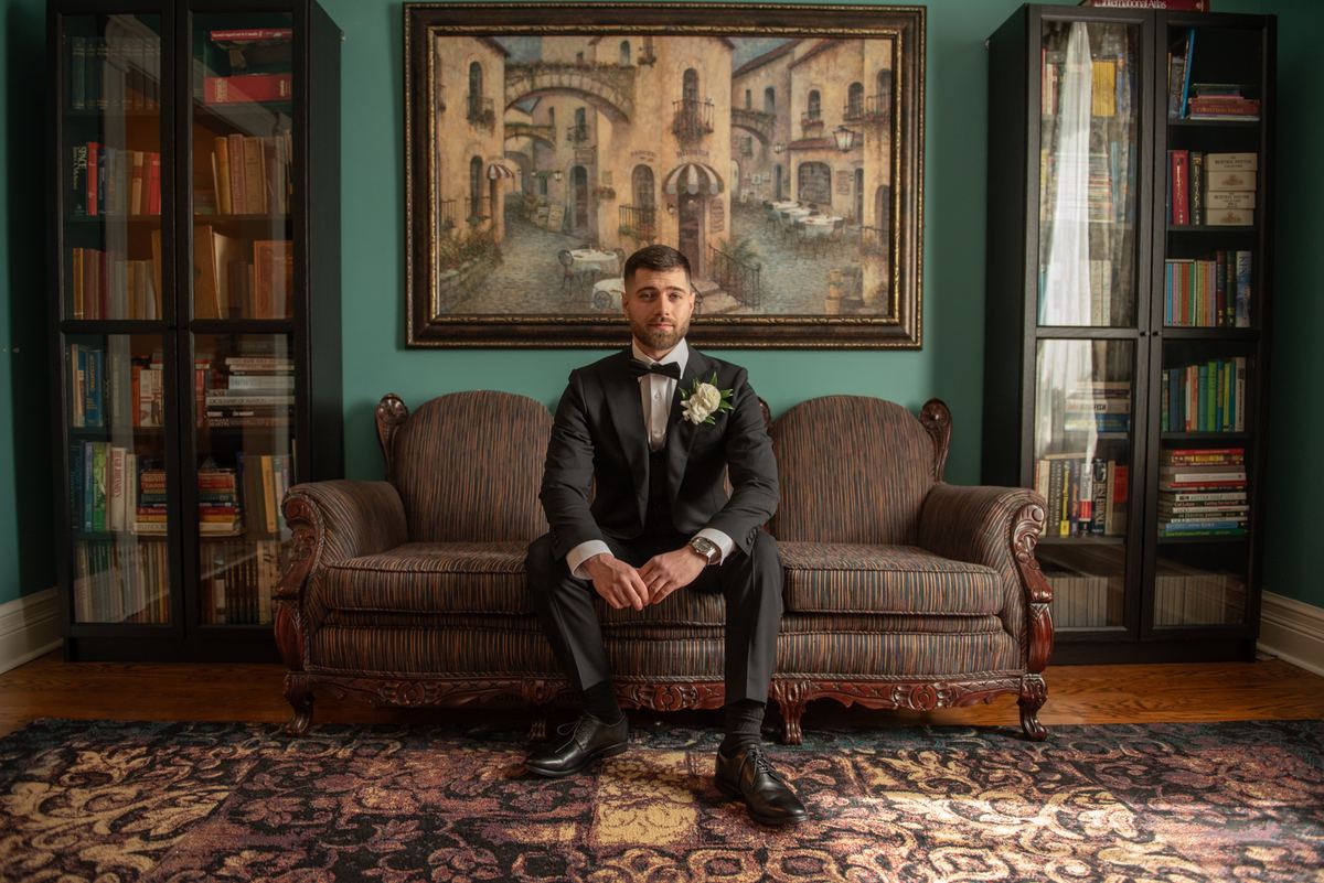 Groom in black tuxedo sits on ornate sofa in richly decorated room with teal walls and book-filled cabinets.