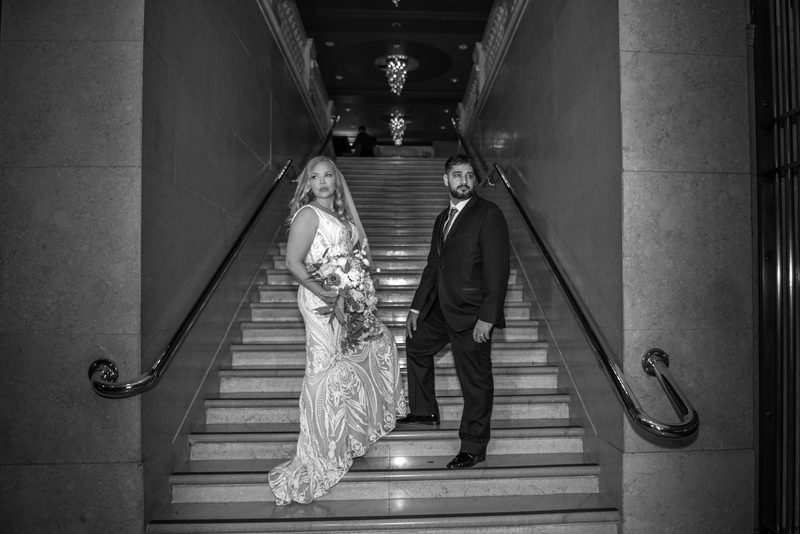 A bride and groom stand on a grand marble staircase, both looking toward the camera in a formal wedding portrait.
