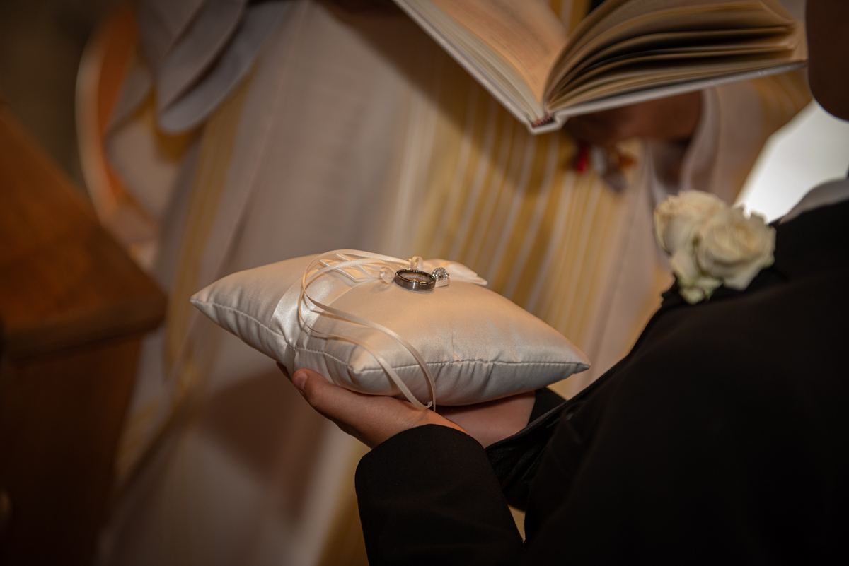 A person in formal attire holds a satin ring pillow with wedding bands, set against an open book and draped fabric.