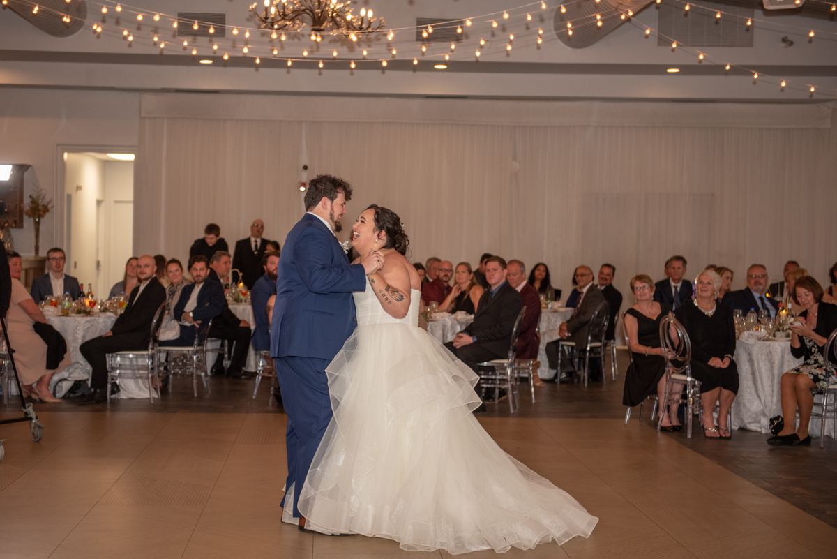 A bride and groom dance joyfully on a wooden floor, surrounded by guests at decorated tables under warm lights.