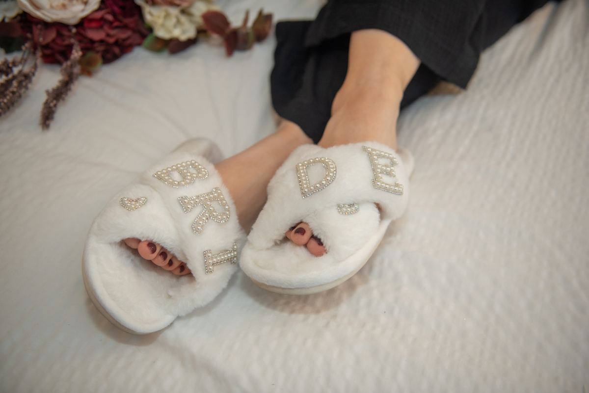 A bride relaxes in white slippers with sparkling 'BRIDE' lettering beside dried flowers on a bed, bathed in soft light.