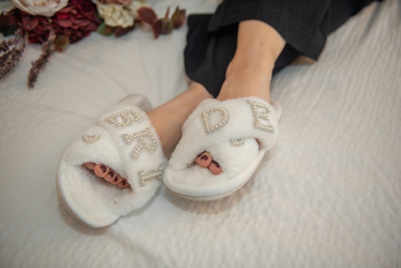 A bride relaxes in white slippers with sparkling 'BRIDE' lettering beside dried flowers on a bed, bathed in soft light.
