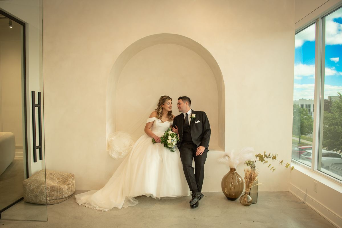 A newlywed couple sits closely in an arched alcove, illuminated by natural light streaming through a window to their right.
