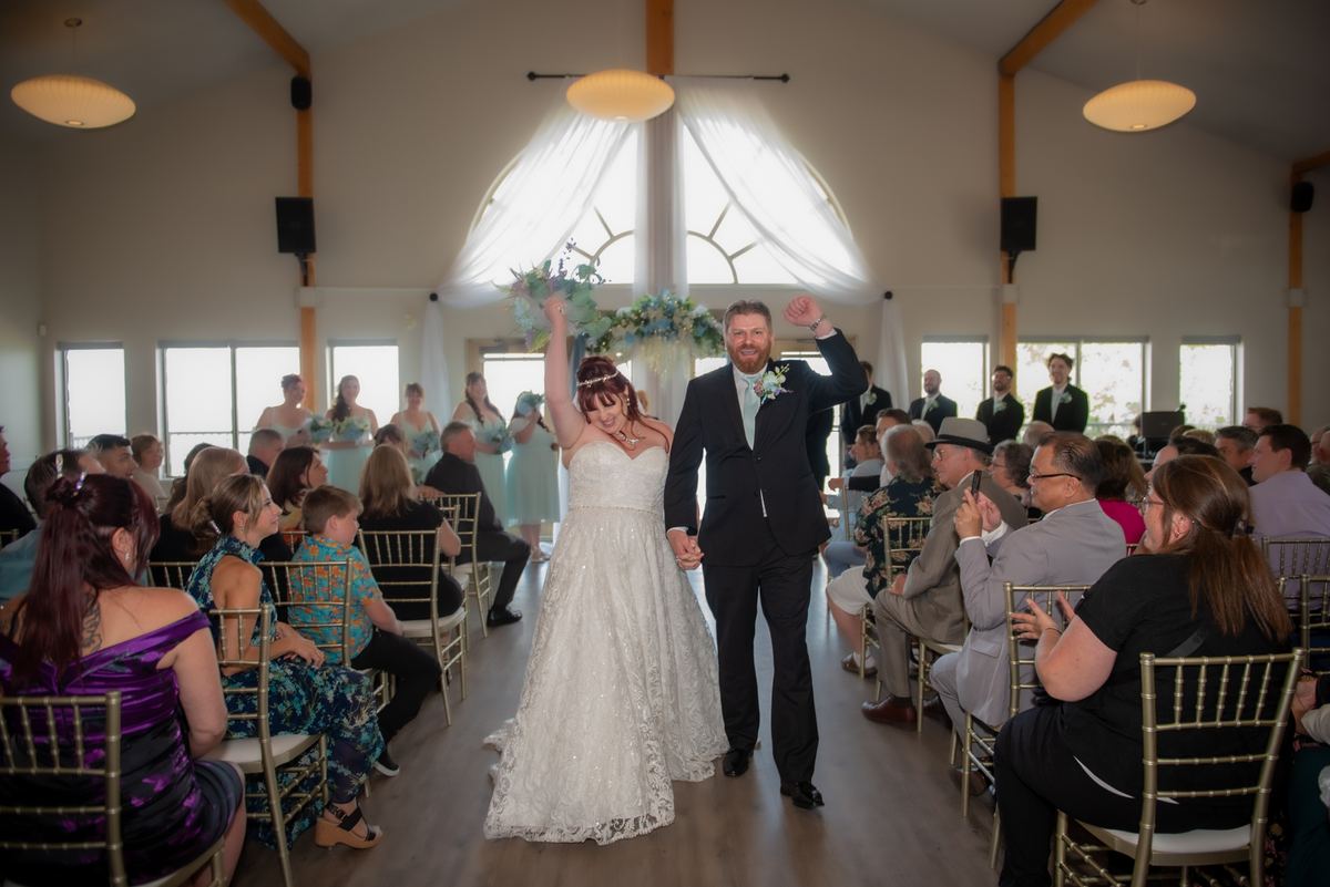 A bride and groom walk down the aisle, arms raised in celebration, illuminated by natural light streaming through windows.