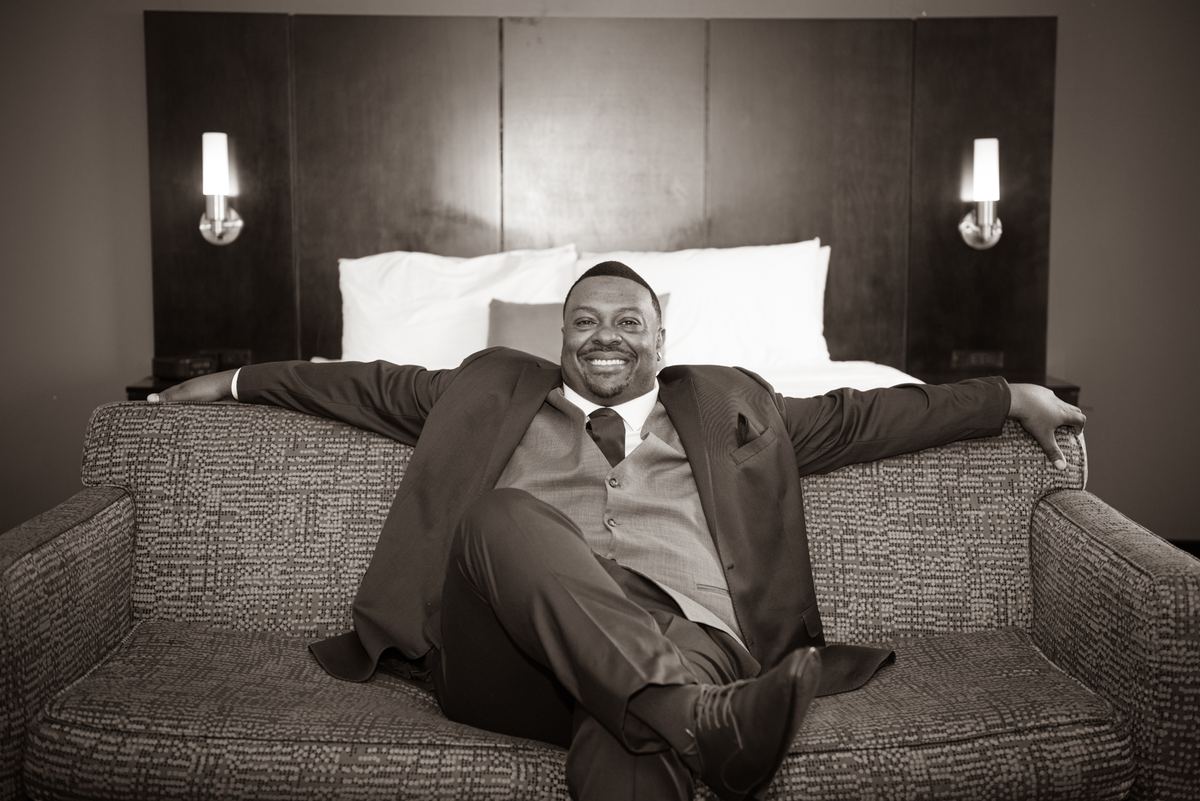 A man in formal wear reclines confidently in an armchair, smiling broadly at the camera in sepia-toned monochrome.