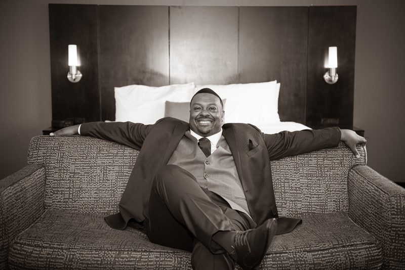 A man in formal wear reclines confidently in an armchair, smiling broadly at the camera in sepia-toned monochrome.