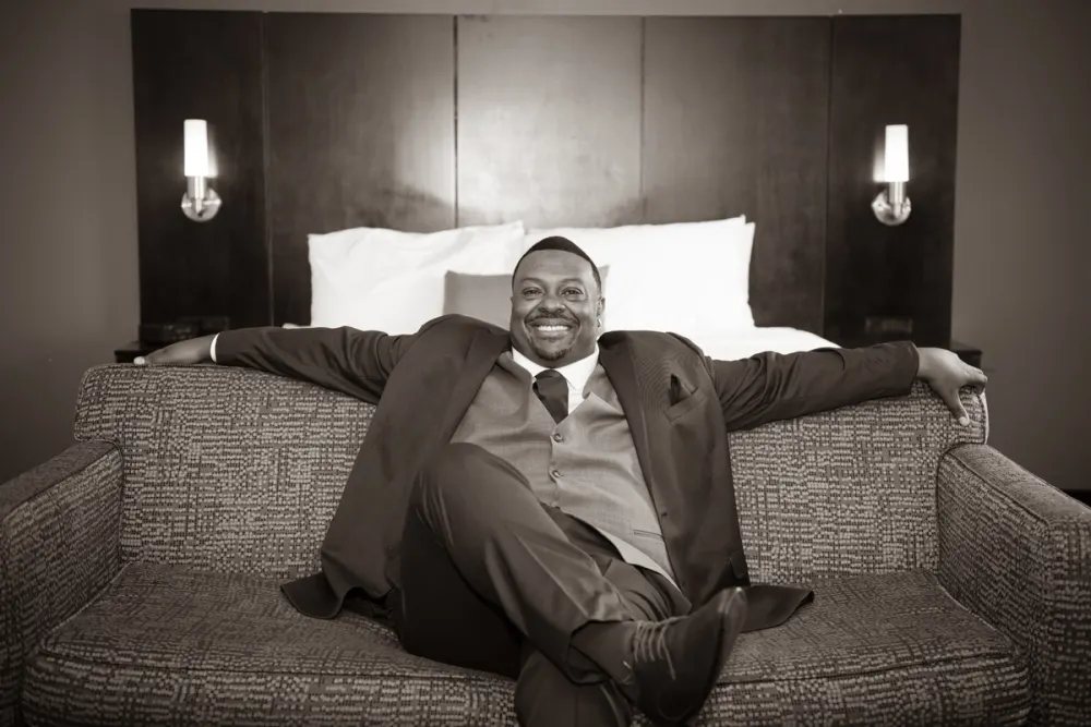 A man in formal wear reclines confidently in an armchair, smiling broadly at the camera in sepia-toned monochrome.