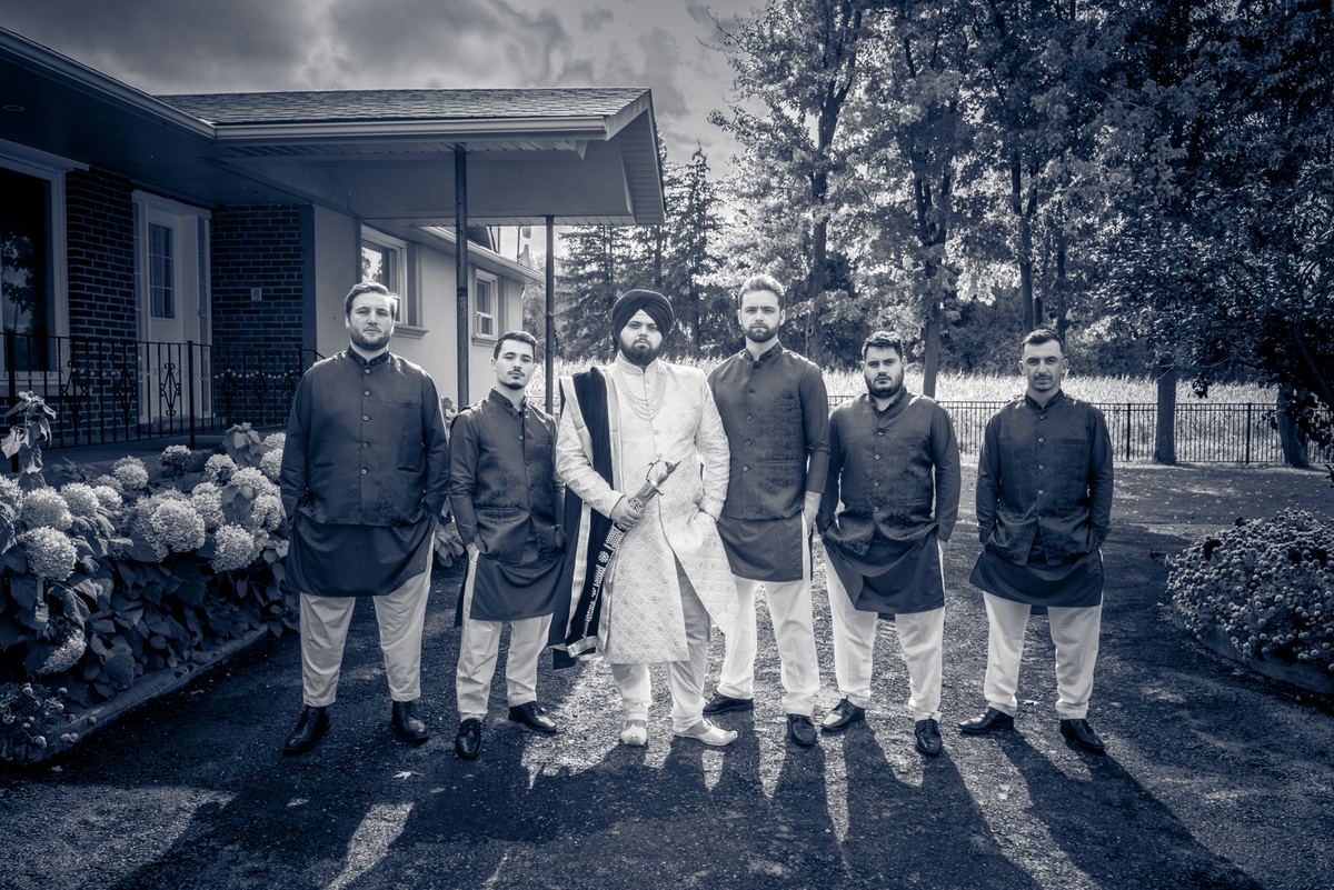 A groom in traditional attire and his groomsmen stand outdoors for pre-wedding photos, framed by trees and a house.