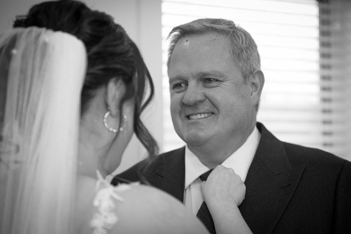 A bride in veil adjusts her father's tie as he smiles warmly, lit by soft natural light through blinds.