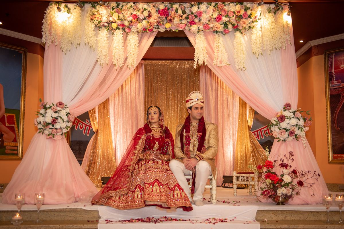 A newlywed couple in traditional attire sits on a decorated stage under a floral canopy, illuminated by warm lights.