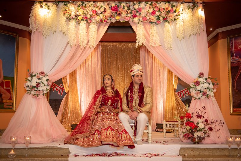 A newlywed couple in traditional attire sits on a decorated stage under a floral canopy, illuminated by warm lights.