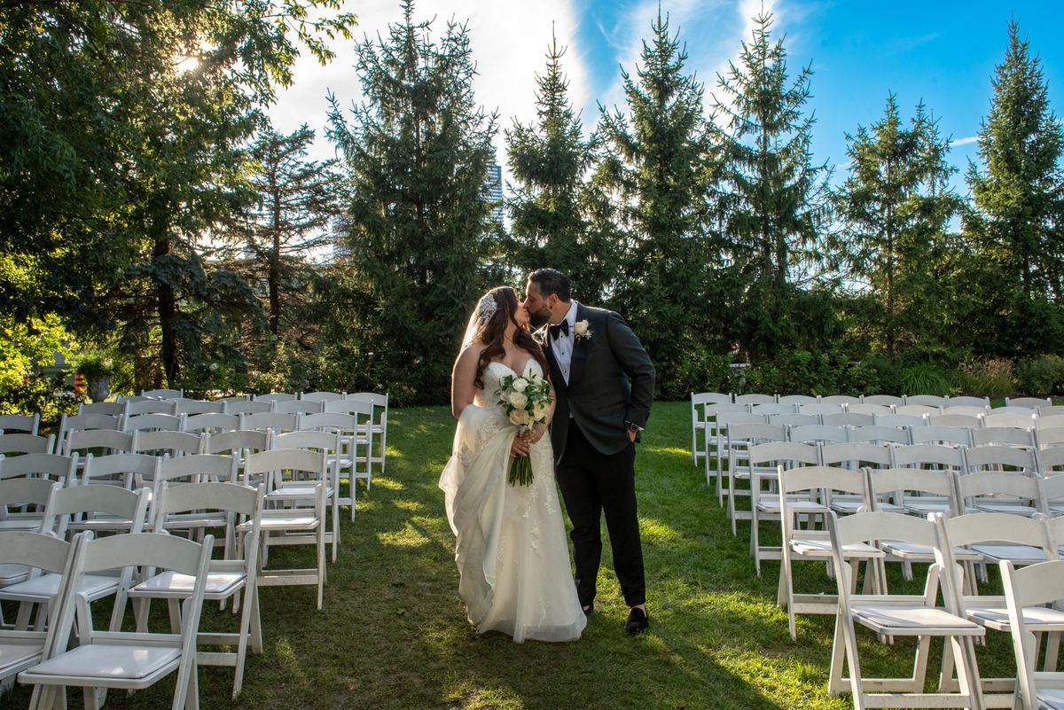 A newlywed couple kisses in a sunlit grass aisle, surrounded by white chairs and evergreen trees casting long shadows.