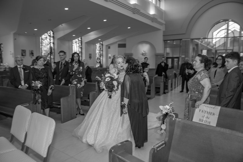 A bride in a sparkling gown walks down the aisle of a modern church, embraced by a woman in a dark robe.