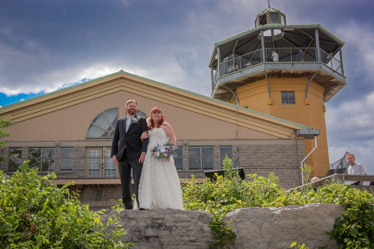 A newlywed couple smiles on a stone ledge in front of a yellow tower, with green foliage and a man in white visible.