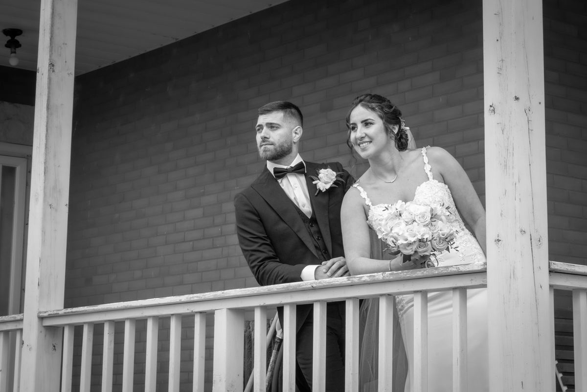 Bride and groom stand on wooden porch railing, gazing out with joy, in soft light highlighting textures of brick wall.