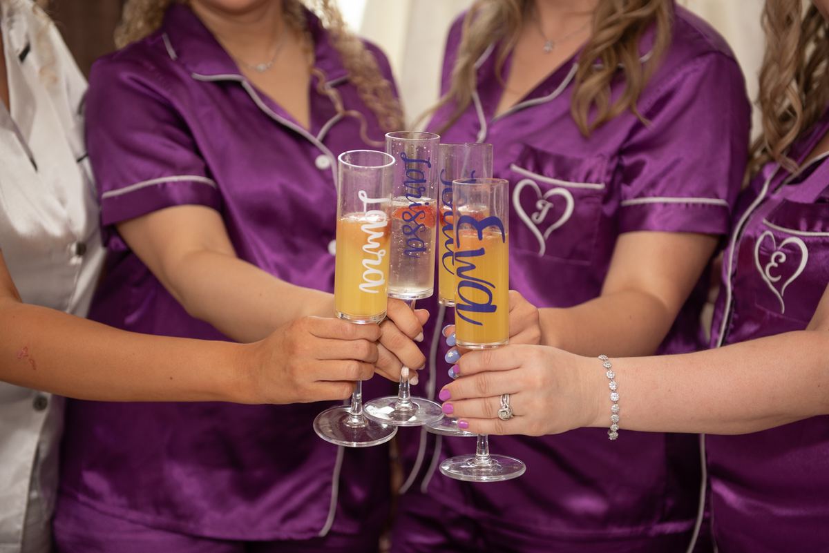 Women in purple pajamas toast with personalized champagne flutes filled with orange-hued drinks, hands clasped together.