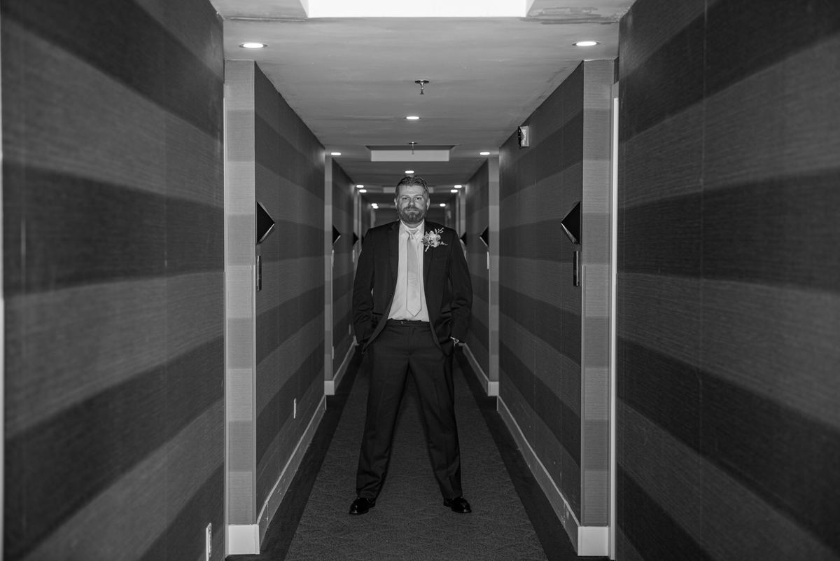 A groom in a suit with a boutonnière stands confidently in a narrow hotel corridor, hands in pockets.