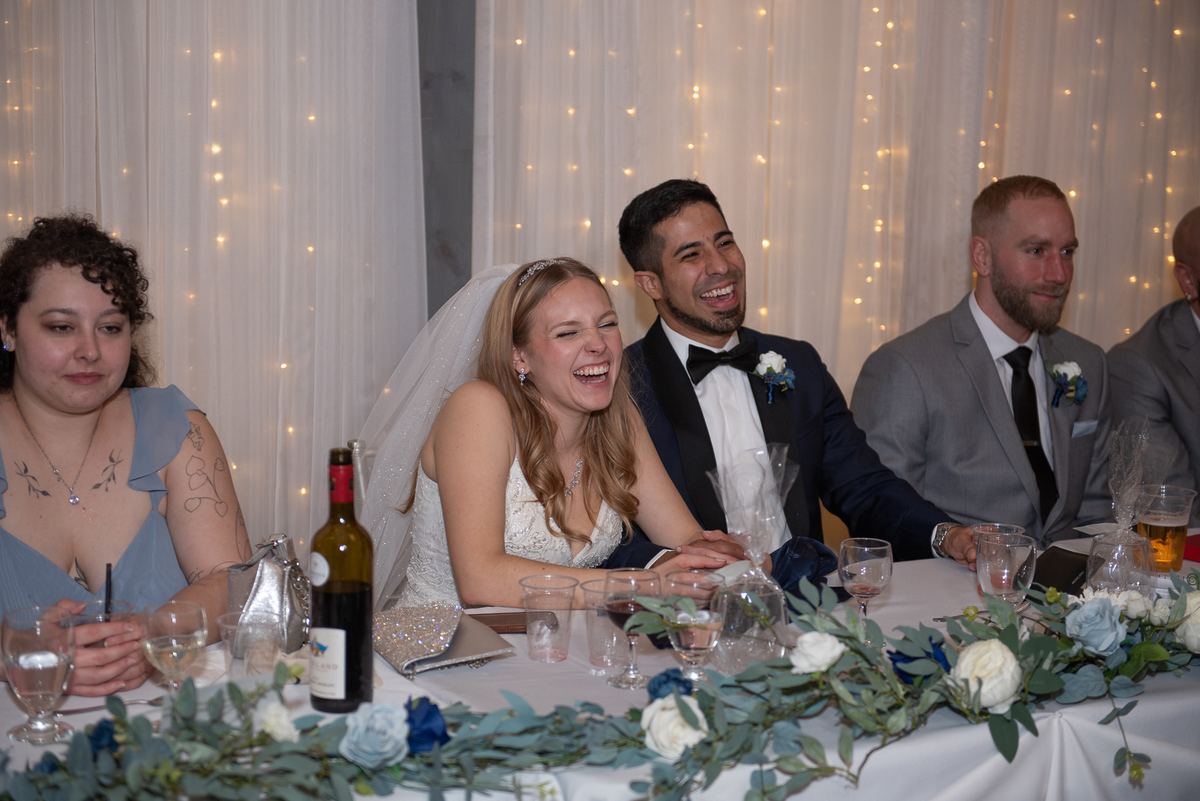 The bride laughs beside her groom at an elegantly set table with floral arrangements and string lights in the background.