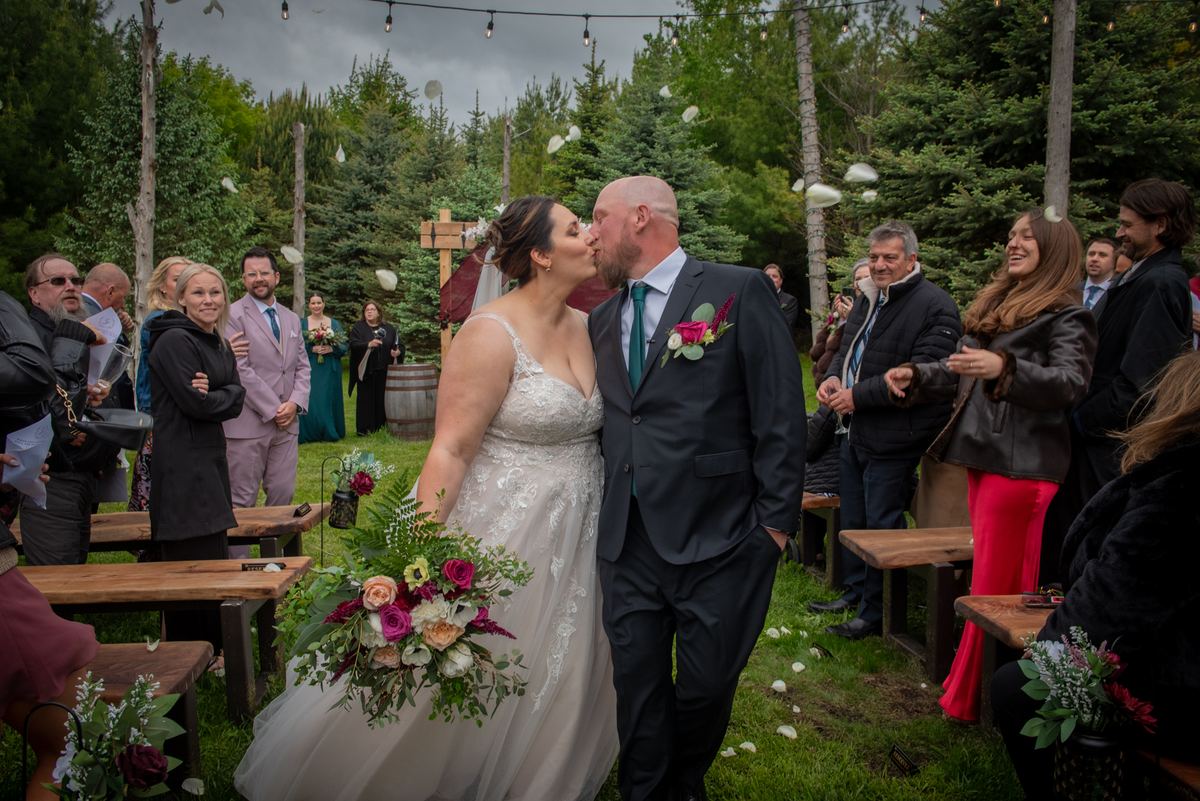 Bride and groom kiss as guests toss petals, surrounded by wooden benches and string lights under an overcast sky.