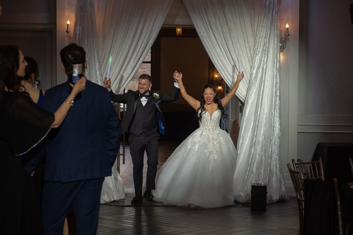 A smiling bride and groom celebrate by raising their arms as guests take photos, illuminated by warm sconce light.