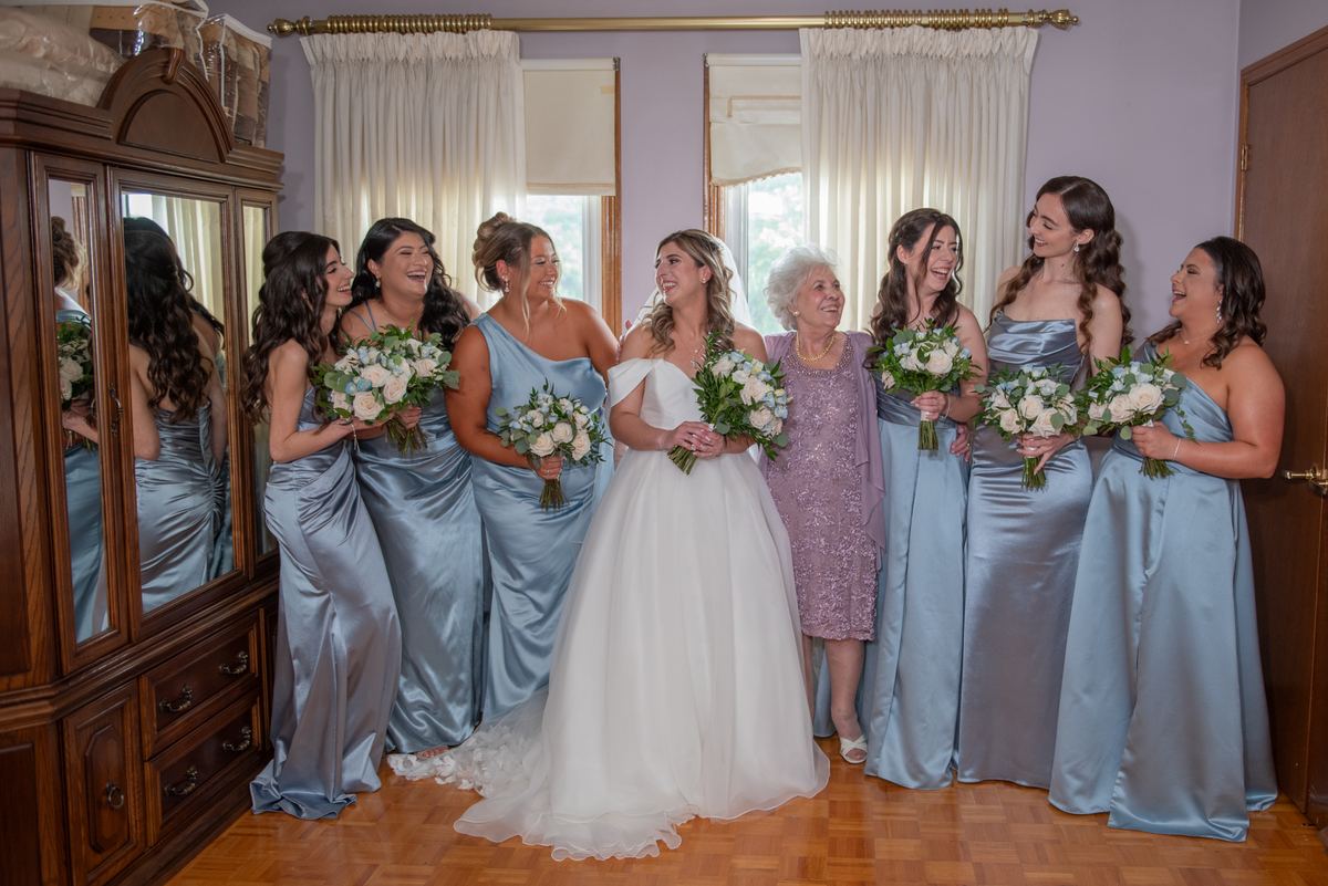 A bride and her bridesmaids in pastel dresses laugh together with bouquets of white and blush flowers.