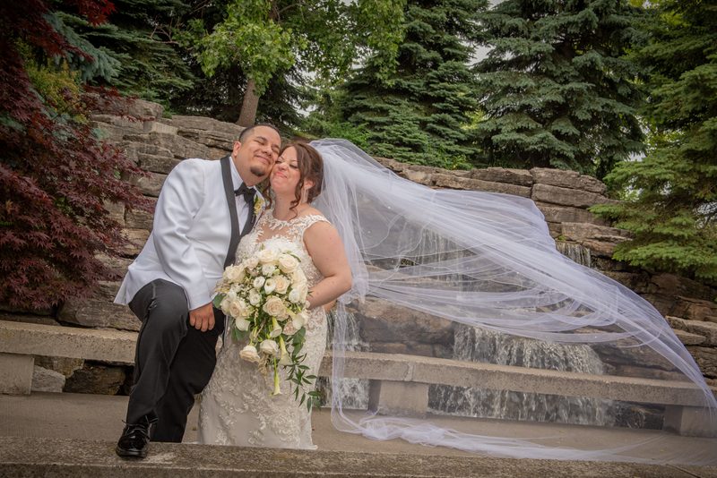 A bride and groom pose on stone steps beside a waterfall, with the bride's veil blowing in the wind.