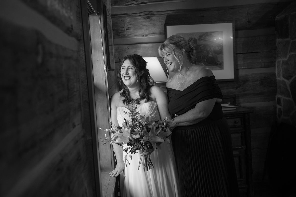 A bride and a woman, likely her mother, share a joyful laugh by a doorway, illuminated by soft light.