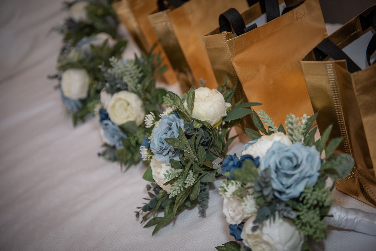 Bouquets of blue and white roses with greenery next to golden gift bags on a white surface, ready for an event.
