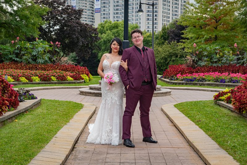A smiling bride and groom stand together in an urban park, surrounded by colorful flowerbeds and tall buildings.